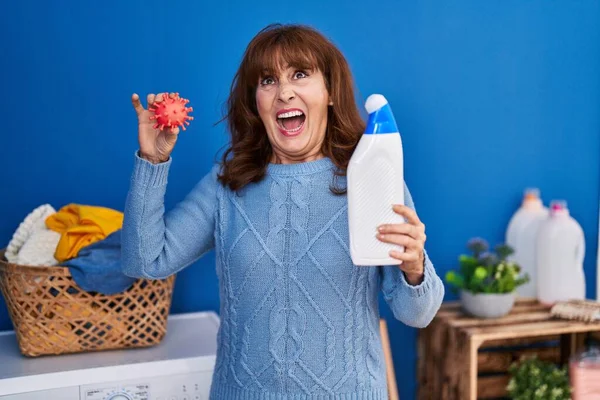 Middle age hispanic woman holding detergent bottle cleaning virus angry and mad screaming frustrated and furious, shouting with anger looking up.