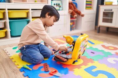 Adorable hispanic boy playing with technician tools toy sitting on floor at kindergarten