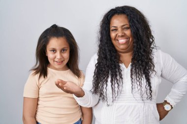 Anne ve küçük kızı beyaz arka planda durup mutlu bir şekilde sunum yapıyor ve avuç içiyle kameraya bakıyor.. 