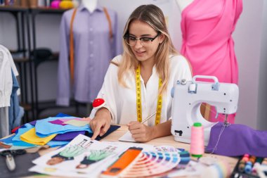 Young blonde woman tailor pointing to clothing design speaking at clothing shop