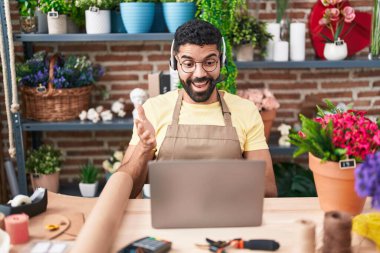 Hispanic man with beard working at florist shop doing video call celebrating achievement with happy smile and winner expression with raised hand 