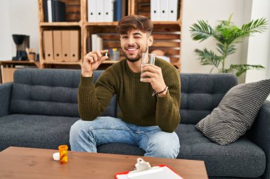 Arab man with beard working on depression holding pills and water winking looking at the camera with sexy expression, cheerful and happy face. 