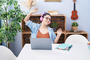 Young caucasian woman sitting on table listening to music and dancing at home