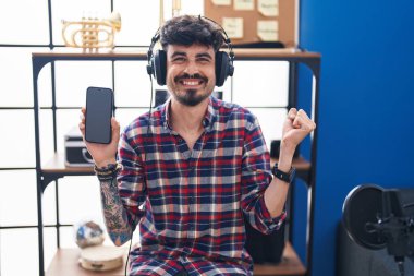 Young hispanic man with beard showing smartphone screen at music studio screaming proud, celebrating victory and success very excited with raised arm 