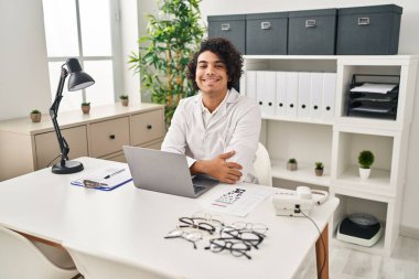Hispanic man with curly hair working at optician office with a happy and cool smile on face. lucky person. 
