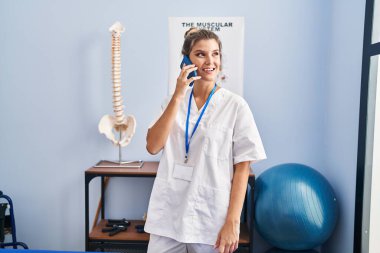 Fizyoterapist üniforması giyen genç sarışın kadın klinikte akıllı telefondan konuşuyor.