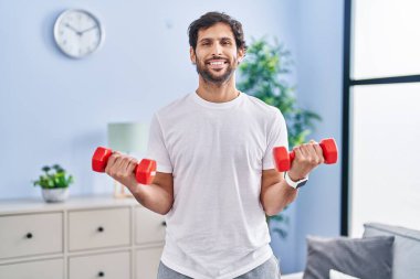 Young hispanic man smiling confident using dumbbells training at home