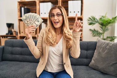 Young blonde therapist woman working at therapy office holding money surprised with an idea or question pointing finger with happy face, number one 