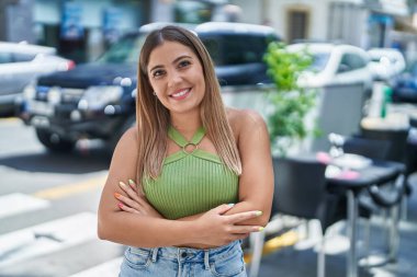 Young beautiful hispanic woman standing with arms crossed gesture at coffee shop terrace