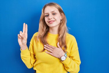 Young caucasian woman standing over blue background smiling swearing with hand on chest and fingers up, making a loyalty promise oath 