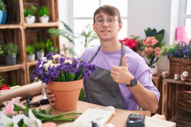 Caucasian blond man working at florist shop doing happy thumbs up gesture with hand. approving expression looking at the camera showing success. 