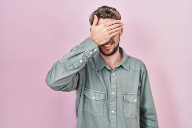 Hispanic man with beard standing over pink background smiling and laughing with hand on face covering eyes for surprise. blind concept. 