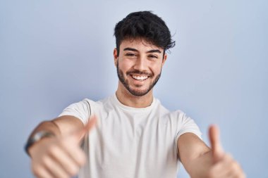 Hispanic man with beard standing over white background approving doing positive gesture with hand, thumbs up smiling and happy for success. winner gesture. 