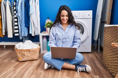 Çamaşır odasında çamaşır makinesi bekleyen genç İspanyol kadın dizüstü bilgisayar kullanıyor.
