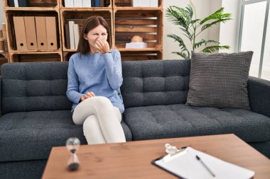 Young brunette woman at consultation office smelling something stinky and disgusting, intolerable smell, holding breath with fingers on nose. bad smell 