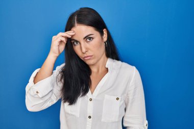 Young hispanic woman standing over blue background worried and stressed about a problem with hand on forehead, nervous and anxious for crisis 