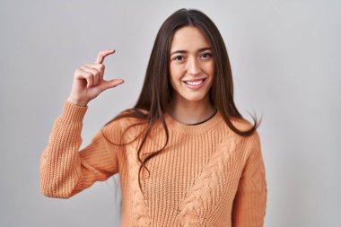 Young brunette woman standing over white background smiling and confident gesturing with hand doing small size sign with fingers looking and the camera. measure concept. 