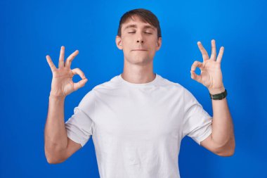 Caucasian blond man standing over blue background relaxed and smiling with eyes closed doing meditation gesture with fingers. yoga concept. 