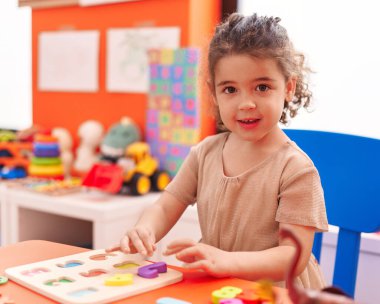 Adorable hispanic girl playing with maths puzzle game sitting on table at kindergarten