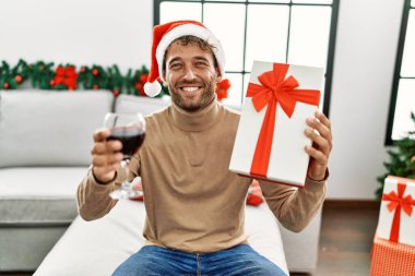 Genç İspanyol adam gülümsüyor, kendine güveniyor, elinde Noel hediyesi tutuyor ve evde şarap içiyor.
