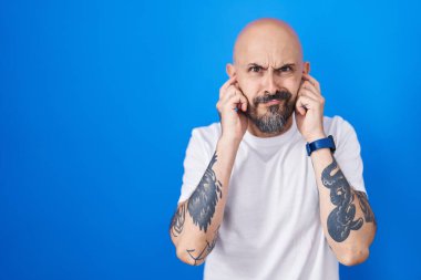 Hispanic man with tattoos standing over blue background covering ears with fingers with annoyed expression for the noise of loud music. deaf concept. 