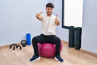 Hispanic man with beard sitting on pilate balls at yoga room approving doing positive gesture with hand, thumbs up smiling and happy for success. winner gesture. 
