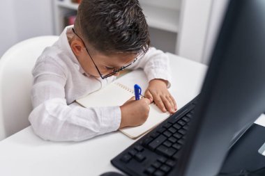 Adorable hispanic boy student using computer writing on notebook at classroom