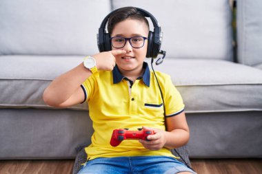 Young hispanic kid playing video game holding controller wearing headphones pointing with hand finger to face and nose, smiling cheerful. beauty concept 