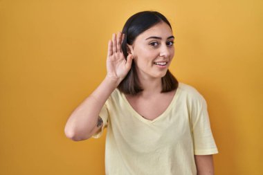 Hispanic girl wearing casual t shirt over yellow background smiling with hand over ear listening an hearing to rumor or gossip. deafness concept. 