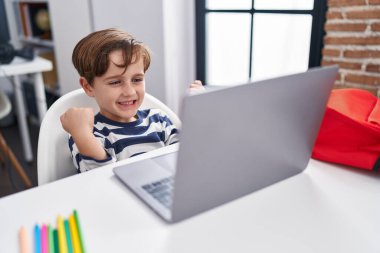 Adorable hispanic boy student using laptop with cheerful expression at classroom