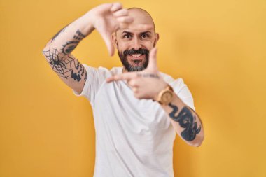 Young hispanic man with tattoos standing over yellow background smiling making frame with hands and fingers with happy face. creativity and photography concept. 