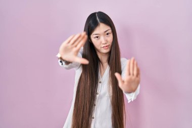 Chinese young woman standing over pink background doing frame using hands palms and fingers, camera perspective 