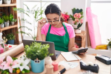 Young hispanic woman florist using laptop and calculator at florist