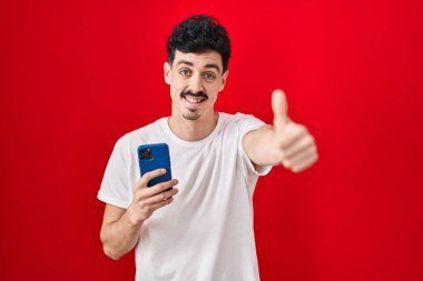 Hispanic man using smartphone over red background approving doing positive gesture with hand, thumbs up smiling and happy for success. winner gesture. 