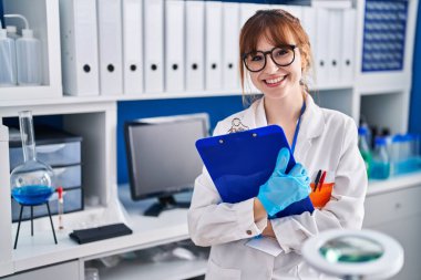 Young woman scientist smiling confident holding clipboard at laboratory