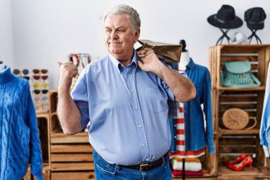 Senior man with grey hair holding shopping bags at retail shop pointing thumb up to the side smiling happy with open mouth 