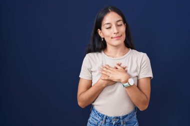 Mavi arka planda duran genç İspanyol kadın elleri göğsünde, gözleri kapalı ve yüzünde minnet dolu bir jest ile gülümsüyor. sağlık kavramı. 