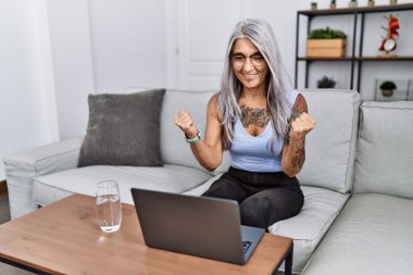 Orta yaşlı, kır saçlı, dizüstü bilgisayar kullanan bir kadın kollarını kaldırıp, gözleri açık bir şekilde başarıyı kutluyor. Kazanan konsept. 