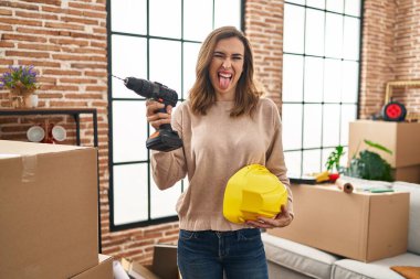 Young woman holding drill at new home sticking tongue out happy with funny expression. 