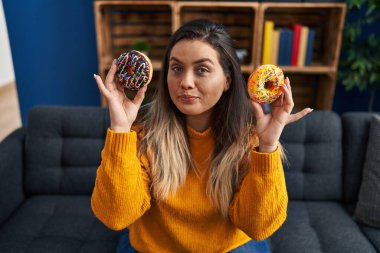 Young hispanic woman eating doughnuts at home smiling looking to the side and staring away thinking. 