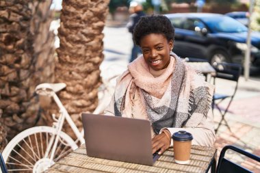 African american woman using laptop drinking coffee sitting on table at coffee shop terrace