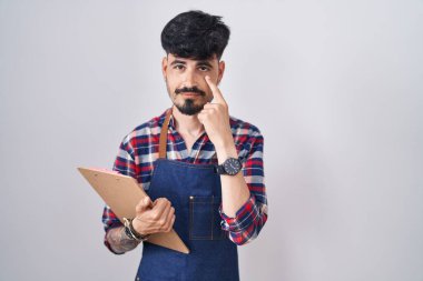 Young hispanic man with beard wearing waiter apron holding clipboard pointing to the eye watching you gesture, suspicious expression 