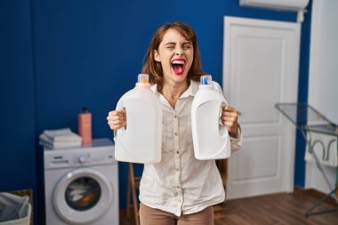 Young beautiful woman holding detergent bottles smiling and laughing hard out loud because funny crazy joke. 