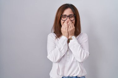 Brunette woman standing over white isolated background laughing and embarrassed giggle covering mouth with hands, gossip and scandal concept 