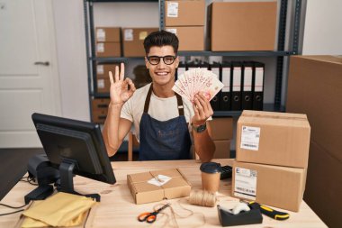 Young hispanic man working at small business ecommerce holding shekels doing ok sign with fingers, smiling friendly gesturing excellent symbol 