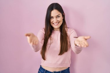 Young brunette woman standing over pink background smiling cheerful offering hands giving assistance and acceptance. 