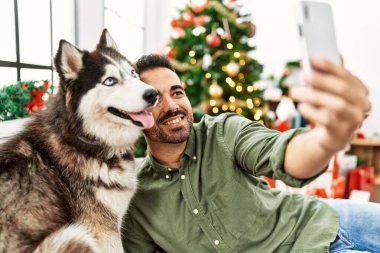 Genç İspanyol adam akıllı telefonuyla selfie çekiyor evdeki yılbaşı ağacında köpekle birlikte kanepede oturuyor.