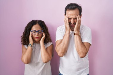 Middle age hispanic couple together over pink background with hand on head, headache because stress. suffering migraine. 