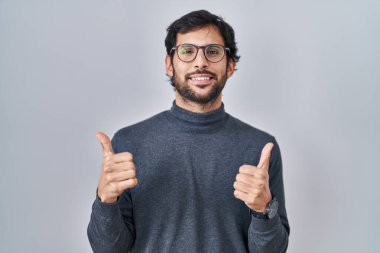 Handsome latin man standing over isolated background success sign doing positive gesture with hand, thumbs up smiling and happy. cheerful expression and winner gesture. 