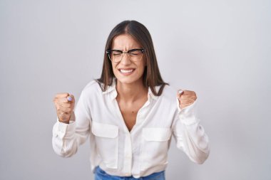 Young brunette woman wearing glasses angry and mad raising fists frustrated and furious while shouting with anger. rage and aggressive concept. 